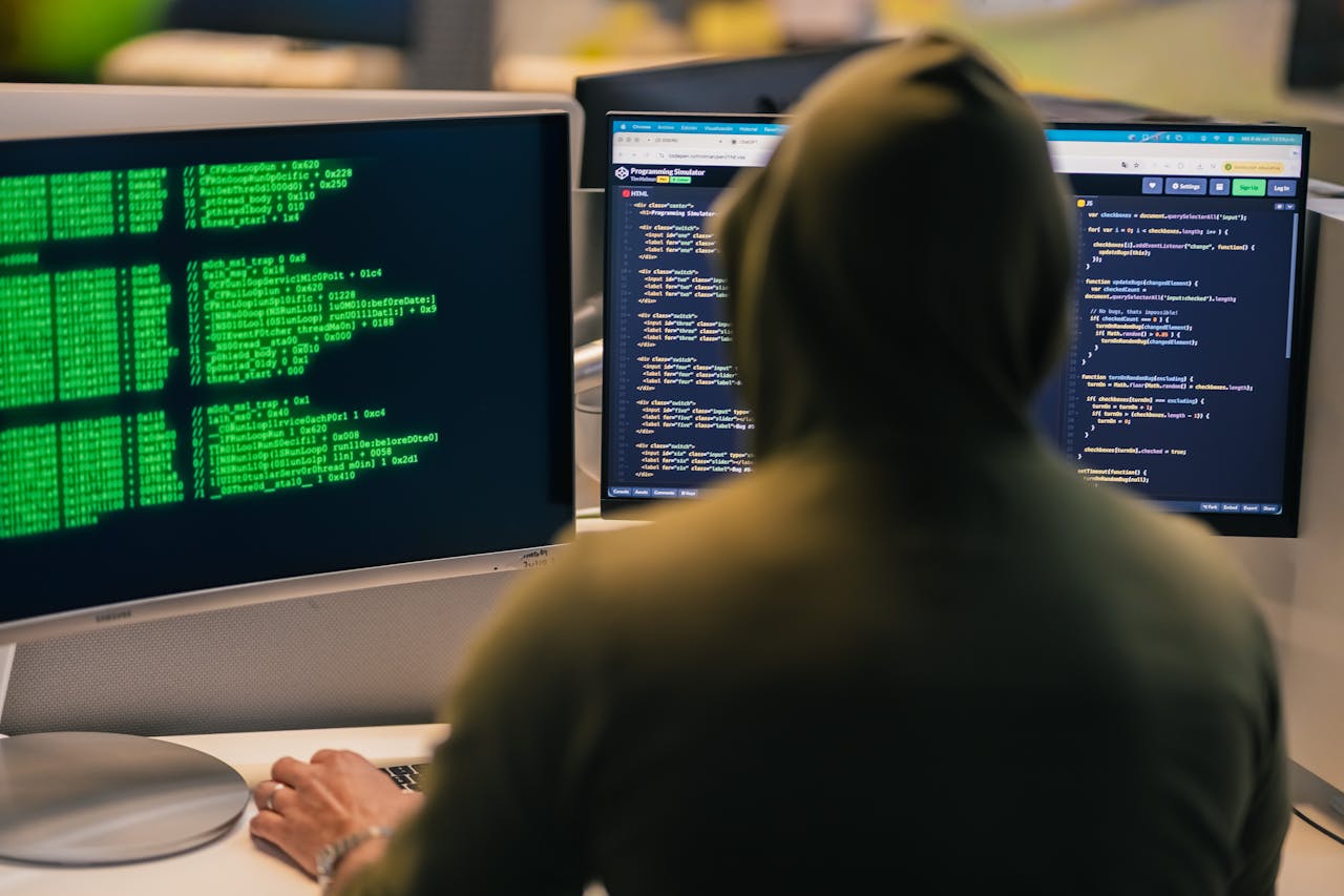 Services A person in a hoodie coding on dual monitors, depicting cybersecurity and hacking themes.